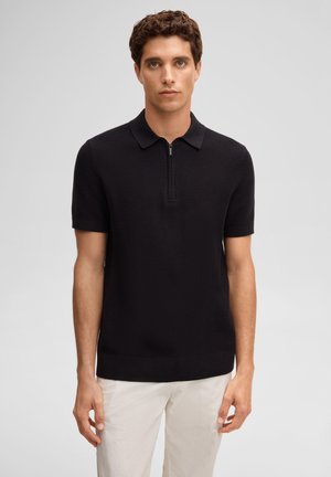 Jonge man met krullend haar die een zwarte korte mouw gebreide polo draagt met een halve rits aan de voorkant en witte broek, staand tegen een effen achtergrond.