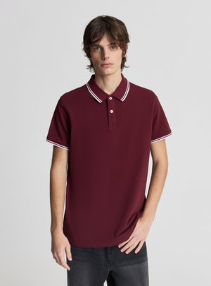 Giovane uomo con capelli castani che indossa una polo color bordeaux con bordi bianchi sul colletto e sulle maniche, in piedi con una mano in tasca.