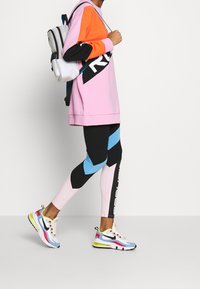 Conjunto atlético que incluye un sudadera de gran tamaño en rosa, naranja y negro, leggings en negro y azul, y zapatillas multicolores. Mochila blanca.