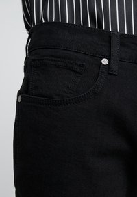 Primer plano de un bolsillo de jeans de mezclilla negros con remaches y un lazo para cinturón, combinado con una camisa de rayas verticales en blanco y negro.