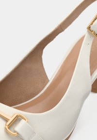 Chaussures à talons blanches avec une finition en cuir lisse, dotées d'une boucle dorée en accent et d'une semelle intérieure beige texturée. La forme du talon est carrée.