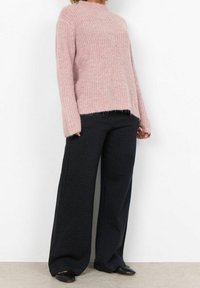 Pull rose clair en maille avec manches longues et col montant, associé à un pantalon large à carreaux foncés et des chaussures plates noires.