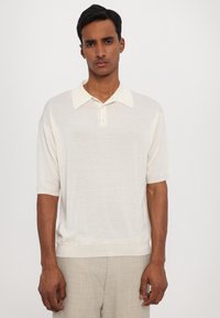 Chemise polo de couleur crème clair, manches courtes, col classique, texture douce, en tissu tricot léger. Associée à un pantalon gris clair texturé.