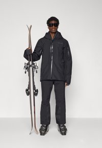 Zwarte waterdichte skijas met een capuchon en ritsen, gecombineerd met bijpassende skibroek. De persoon houdt twee ski's met bindingen vast.