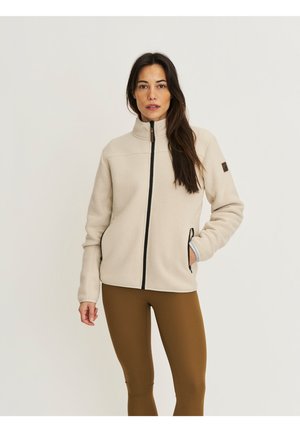 Woman with long dark hair wearing a beige zip-up fleece jacket and brown leggings, standing against a plain light background.