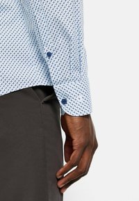 Hellblaues Hemd mit geometrischem Muster, ausgestattet mit zwei Knöpfen an den Manschetten und kontrastierenden dunkelblauen Akzenten. Weiche Stofftextur.