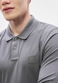 Graues Polohemd mit Kragen, zwei Knöpfen und einem quadratischen Logo-Patch auf der linken Brust. Der Stoff hat eine strukturierte, gestrickte Oberfläche.