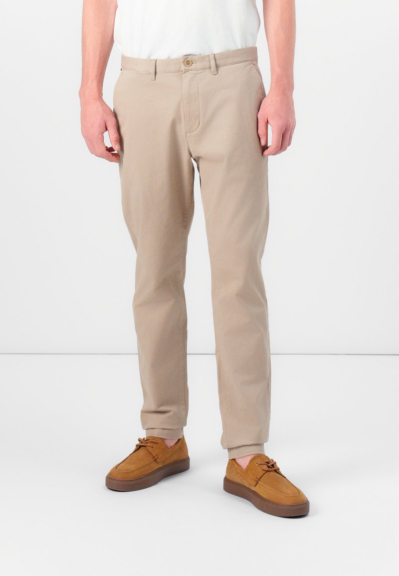 Beige bomulds chinos med lige pasform, lynlås foran og knaplukning. Sammen med brune ruskinds-sko med snørebåndsdesign.