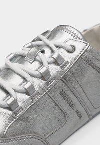 Ασημί αθλητικό παπούτσι από υφή δέρματος με λευκά κορδόνια και μεταλλικά ρουθούνια, που δείχνει το όνομα της μάρκας "MICHAEL KORS" στο πλάι.