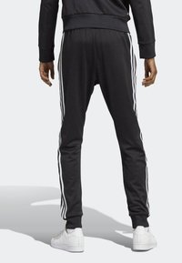 Pantalons de sport noirs avec des bandes blanches sur les côtés, taille élastique et poignet côtelé, présentés de dos avec une main sur la hanche.
