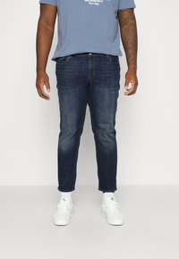 Jeans de mezclilla azul oscuro con corte slim y ligero desgaste, combinados con zapatillas blancas y calcetines blancos hasta el tobillo. Diseño casual y moderno.