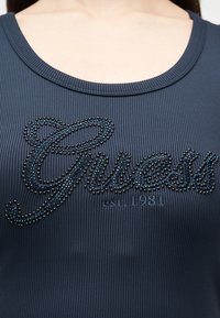 Gros plan d'un haut côtelé bleu foncé avec l'inscription "Guess est. 1981" en strass noirs sur la poitrine.