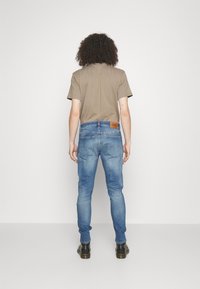 Jasnobrązowy t-shirt w połączeniu z niebieskimi dżinsami z denimu. Dżinsy mają skórzaną metkę z tyłu oraz dwie tylne kieszenie. Czarne buty z gumową podeszwą.