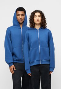 Μπλε φούτερ με φερμουάρ, άνετη εφαρμογή, μεγάλες μπροστινές τσέπες και επενδεδυμένη κουκούλα, κατασκευασμένο από μαλακό, υφασμάτινο ύφασμα με υφή. Μαύρο παντελόνι.