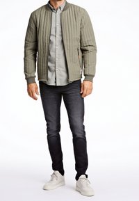Giacca bomber trapuntata verde oliva con polsini a costine, abbinata a una camicia button-up grigia chiara e jeans scuri, completata da sneakers bianche.