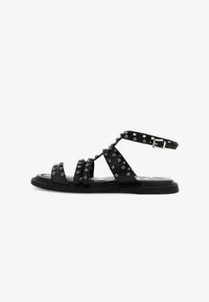 Musta litteä sandaali, jossa useita hihnoja koristeltu pienillä metallinisillä niiteillä ja säädettävä nilkkalukko.