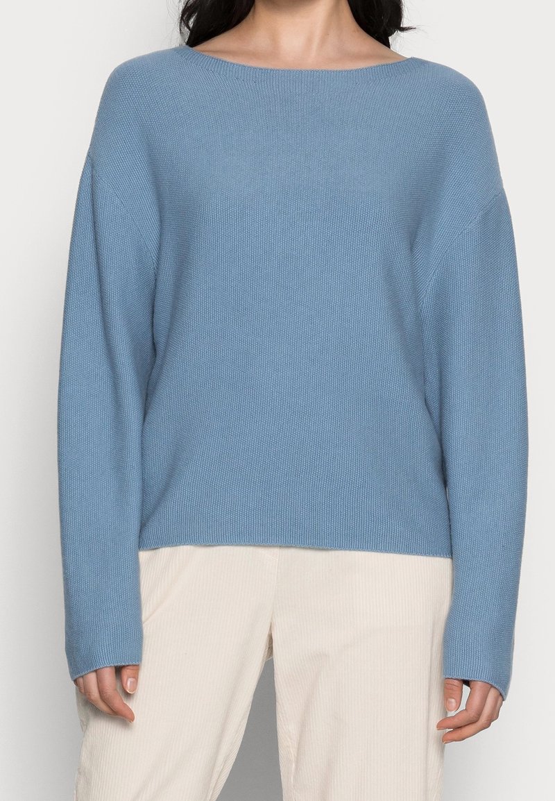 Kobieta ubrana w luźny, jasnoniebieski sweter dzianinowy z długim rękawem i kremowe spodnie, pokazana od ramion do bioder.