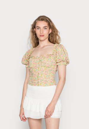 Top floral cropped com mangas bufantes, apresentando uma impressão colorida de flores rosas e azuis em fundo amarelo. Combinado com uma saia branca com babados.