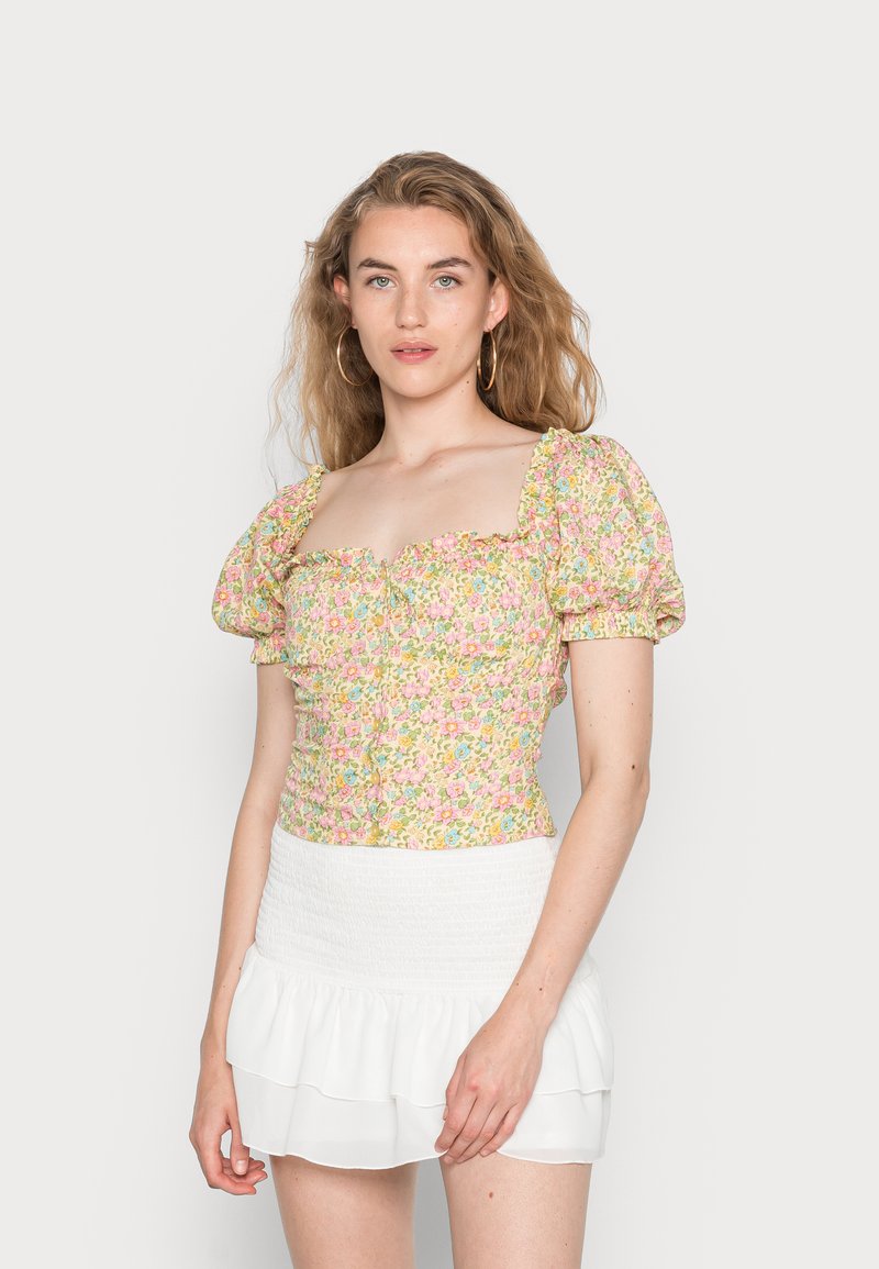 Top floral cropped com mangas bufantes, apresentando uma impressão colorida de flores rosas e azuis em fundo amarelo. Combinado com uma saia branca com babados.