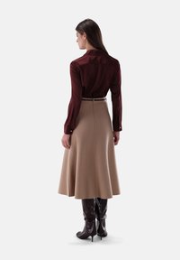 Woman standing with back turned, wearing a burgundy long-sleeve blouse, beige midi skirt, and dark brown knee-high heeled boots.