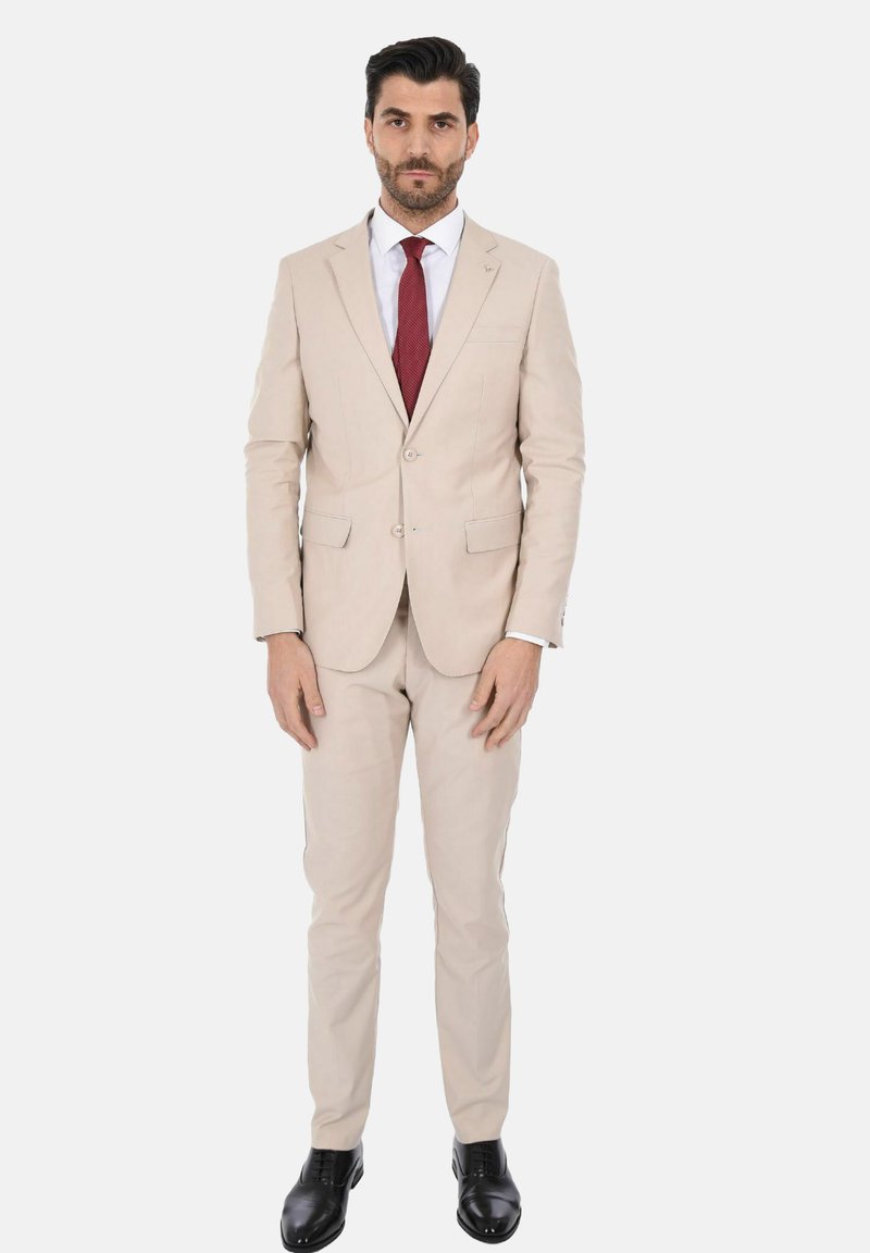 Giacca e pantaloni beige, taglio sartoriale, con due bottoni. Camicia bianca e cravatta rossa. Scarpe eleganti nere, texture liscia.