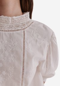 Blusa bianca con delicata ricamo floreale e raffinato bordo in pizzo sul collo alto. Presenta dettagli a pieghe e maniche a sbuffo.
