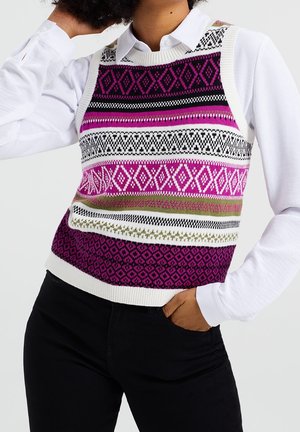 Persoon draagt een wit overhemd onder een mouwloos gebreid vest met paarse, zwarte, witte en groene geometrische patronen, hand in de zak.