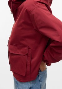Vue de profil d'une personne portant une veste à capuche rouge avec une grande poche à rabat et un jean bleu, main dans la poche arrière.