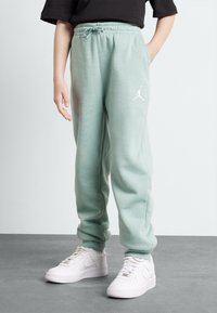 Person som bär mintgröna joggers med dragsko och vit logotyp, kombinerat med vita sneakers, stående på grått golv mot vit vägg.