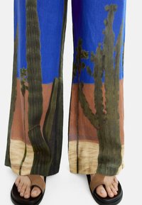 Szerokie spodnie w intensywnym niebieskim kolorze z nadrukiem krajobrazu z kaktusami w zielonych i ziemistych odcieniach. Uzupełnione ciemnobrązowymi klapkami.