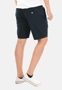 Dark blue shorts made of cotton blend, featuring side pockets and buttoned back pockets. Worn with white sneakers and black socks.