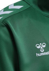 T-shirt de sport vert en tissu lisse, avec des rayures en chevron blanches sur les épaules et un logo blanc sur le bas de l'avant.