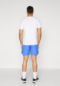 Man standing facing away wearing a white short-sleeve shirt, blue shorts with side pockets, white socks, and black athletic shoes.