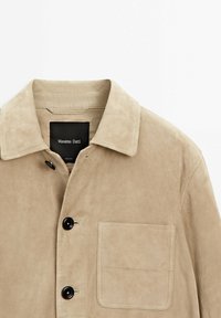 Veste en daim beige avec des boutons noirs, une poche poitrine et un col, étiquetée Massimo Dutti.