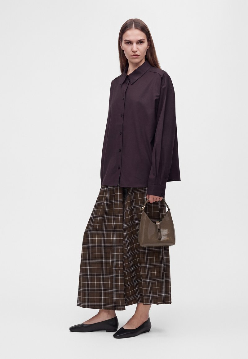 Femme portant une chemise ample boutonnée foncée, un pantalon large marron à carreaux, des ballerines noires, tenant un petit sac à main marron sur un fond uni.