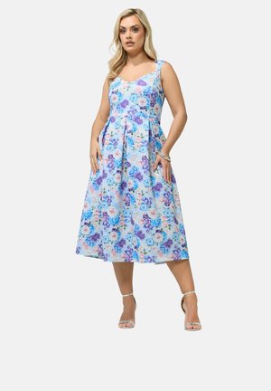 Femme portant une robe sans manches à fleurs bleues et violettes, longueur genou avec des plis, debout les mains dans les poches, chaussée de talons argentés et portant un bracelet.