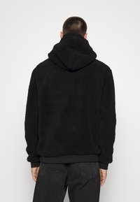 Svart, teksturert fleecehetta med stor hette og elastiske mansjetter; kombinert med mørke jeans, sett fra baksiden mot en hvit bakgrunn.
