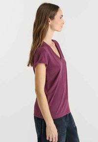 Vrouw met lang bruin haar staat zijwaarts gekeerd en draagt een paars V-hals T-shirt met korte mouwen en donkere broek tegen een effen achtergrond.