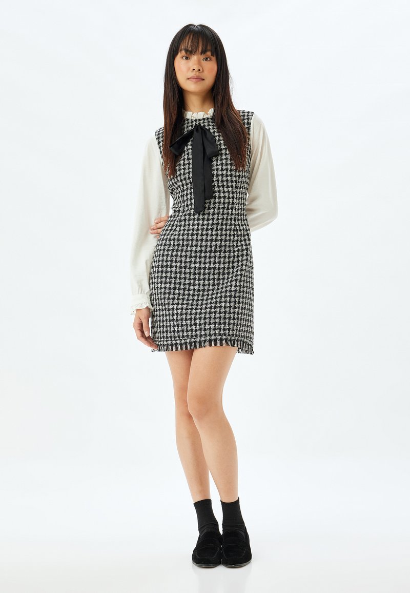 Sort-hvid houndstooth-kjole med frynsekanter, stylet over en cremefarvet bluse med flæsekrave og sort sløjfe. Sorte strømper og sko.