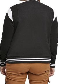 Svart sweatshirt med vita axeldetaljer, ribbad krage och manschetter. Har horisontella vita ränder vid nederkanten. Mjuk textur.