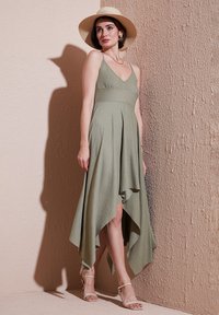 Green, flowy, knee-length dress with a high-low hem, spaghetti straps, and a textured fabric. Worn with a wide-brimmed straw hat and sandals.