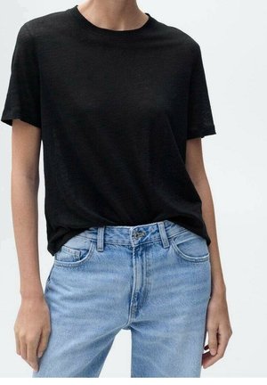 Person trägt ein schlichtes schwarzes kurzärmeliges T-Shirt, das in hellblaue, hoch taillierte Jeans aus Denim gesteckt ist, vor einem schlichten weißen Hintergrund.