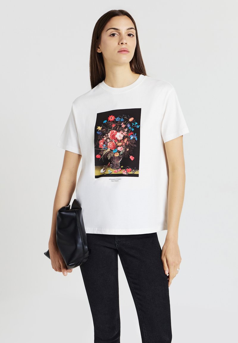 Donna con lunghi capelli scuri, vestita con una t-shirt bianca con una stampa floreale colorata, pantaloni neri e tiene in mano una borsa a pochette nera.