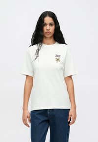 Biela bavlnená tričko s krátkymi rukávmi, na ľavej strane hrudníka s vyšitým zeleným a žltým dizajnom. Voľný strih.