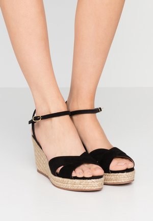 Feet wearing black suede wedge sandals with ankle straps and woven rope platform heels on a light gray floor.