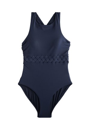 Kobieta z długimi kręconymi włosami w czarnym jednoczęściowym kostiumie kąpielowym stoi na plaży z falami w tle.