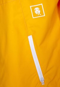 Żółta kurtka z białą kieszenią na zamek oraz kwadratowym logo z wizerunkiem twarzy. Tkanina wygląda na gładką z delikatnym połyskiem.