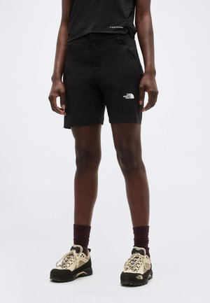 Άτομο που φοράει μαύρο σορτς υπαίθρου με το λογότυπο The North Face, συνδυασμένο με μπεζ παπούτσια πεζοπορίας και σκούρες κάλτσες, στέκεται σε απλό φόντο.