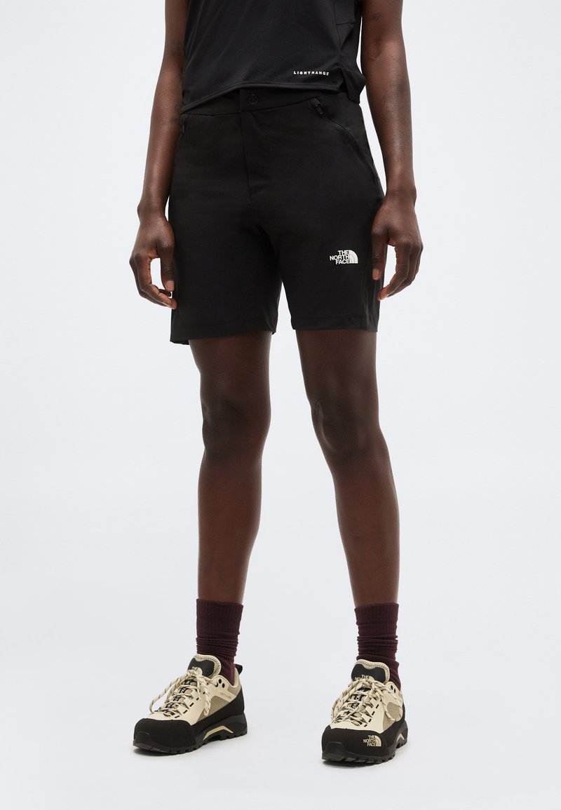 Henkilö, joka käyttää mustia ulkoiluhousuja, joissa on The North Face -logo, yhdistettynä beigenvärisiin vaelluskenkiin ja tummiin sukkiin, seisoo yksinkertaisella taustalla.