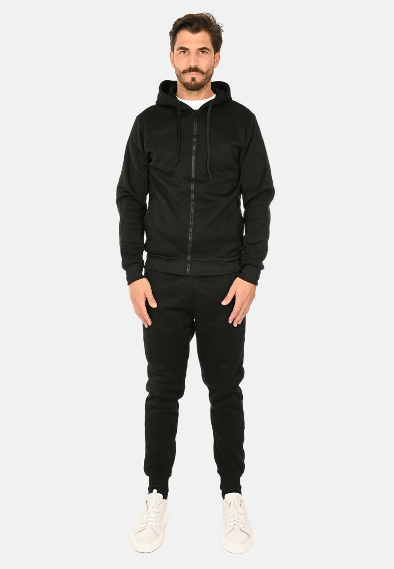 Felpa nera con zip e cappuccio, abbinata a pantaloni jogger neri. Presenta polsini e vita a coste, indossata con sneakers bianche.